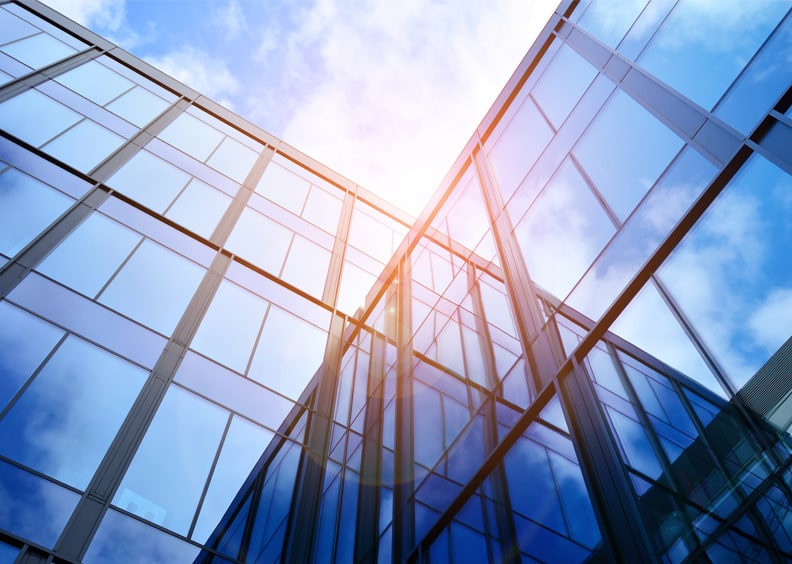 A low-angle view of a modern glass building with sunlight reflecting off its surface, set against a clear blue sky.
