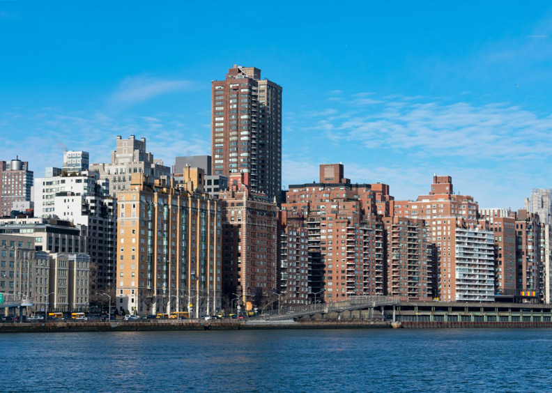 A city skyline with multiple mid- and high-rise buildings along a waterfront under a clear blue sky.